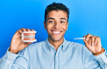 Young man with perfect smile holding a dental model with braces in one hand and an invisible aligner in the other hand.